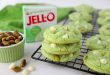 Stack of soft pistachio pudding cookies with white chocolate chips and chopped pistachios on a cooling rack, next to a box of pistachio pudding and a ramekin of whole pistachios