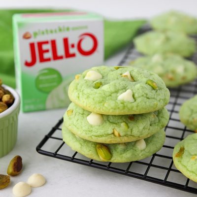 Stack of soft pistachio pudding cookies with white chocolate chips and chopped pistachios on a cooling rack, next to a box of pistachio pudding and a ramekin of whole pistachios