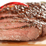 Juicy, sliced carne asada with a charred crust, garnished with red chili peppers on a wooden cutting board.