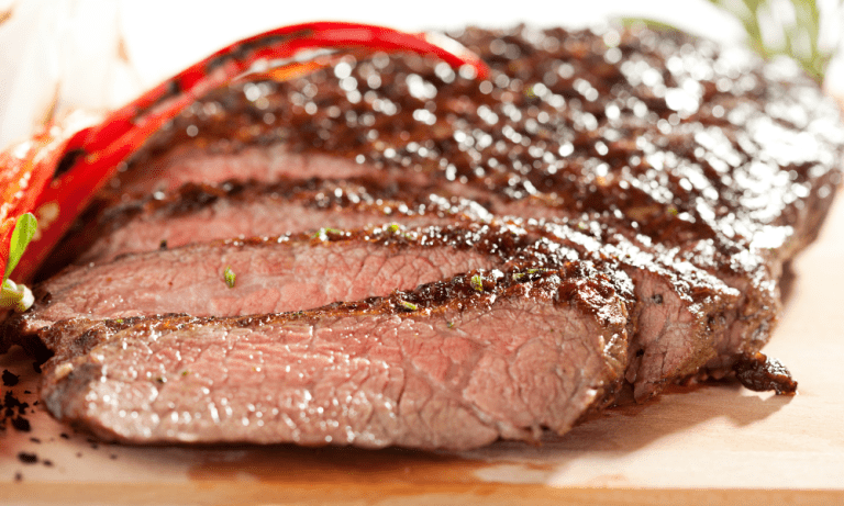 Juicy, sliced carne asada with a charred crust, garnished with red chili peppers on a wooden cutting board.