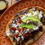 A classic Mexican huarache adorned with savory beef, creamy cheese, fresh avocado slices, and vibrant red and green salsas, served alongside small bowls of salsa for dipping.