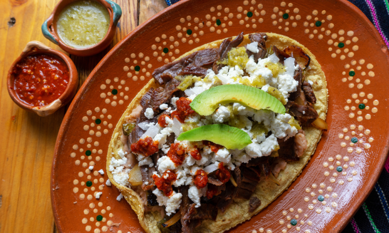 A classic Mexican huarache adorned with savory beef, creamy cheese, fresh avocado slices, and vibrant red and green salsas, served alongside small bowls of salsa for dipping.