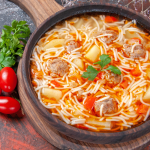 Top view of beef soup with meatballs, potatoes, and noodles, surrounded by fresh vegetables and spices on a dark table.