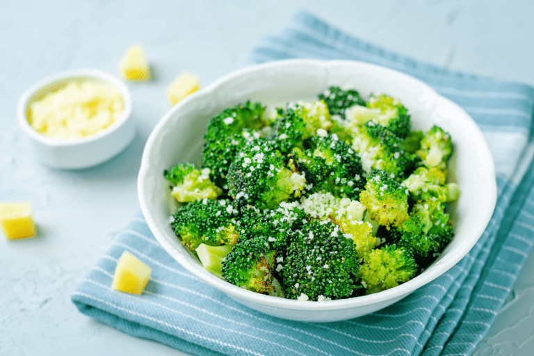 A simple white bowl packed with roasted broccoli, topped generously with grated cheese. The soft hues of the background make the green florets pop
