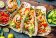 A closer look at shrimp cradled in soft tortillas and topped with chopped lettuce, peppers, and herbs. The accompanying bowls of pico de gallo and diced avocado invite you to customize each bite.