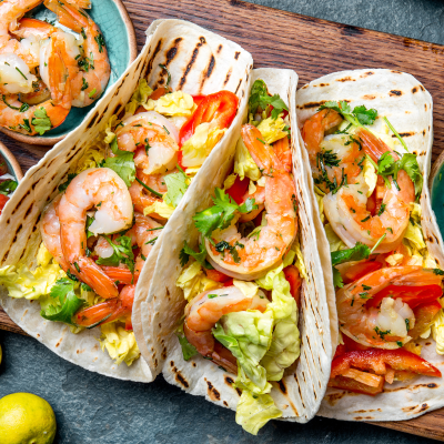 A closer look at shrimp cradled in soft tortillas and topped with chopped lettuce, peppers, and herbs. The accompanying bowls of pico de gallo and diced avocado invite you to customize each bite.