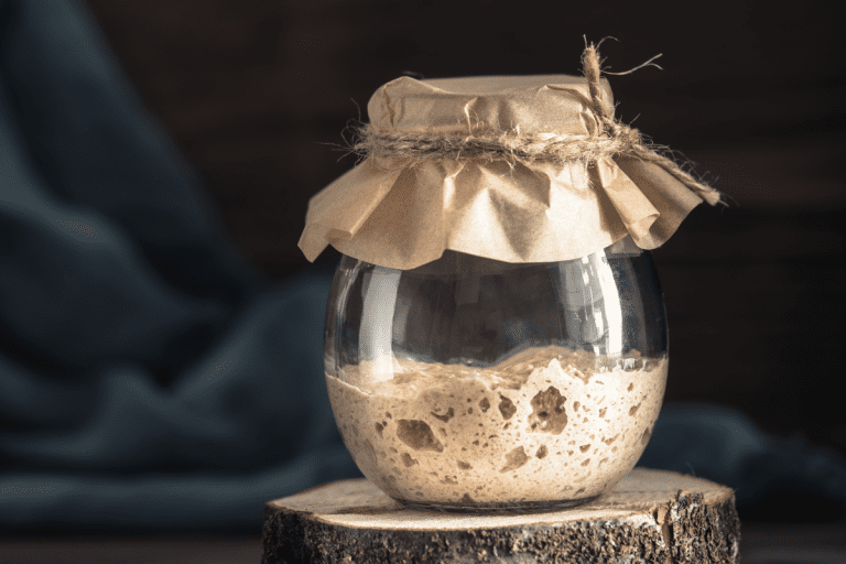 A partially filled glass jar of Sourdough Discard Recipes, starter, covered with kraft paper and tied with rustic twine, resting on a wood slice.