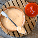 A single shot of a terracotta bowl filled with pink Yum Yum Sauce on a bamboo trivet, paired with a spoon and a small glass of ketchup on the side.