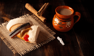 Traditional Mexican Rosca de Reyes slice with colorful candied fruit atop soft, sugar-dusted bread, paired with a rustic clay mug of hot chocolate and cinnamon sticks on a dark wooden table. A small baby figurine symbolizes the cultural tradition of Epiphany celebration.