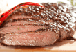 Juicy, sliced carne asada with a charred crust, garnished with red chili peppers on a wooden cutting board.
