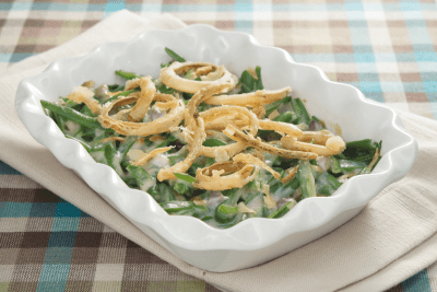 Close-up of a baked green bean casserole in a white dish, topped with golden-brown fried onions on a plaid tablecloth.