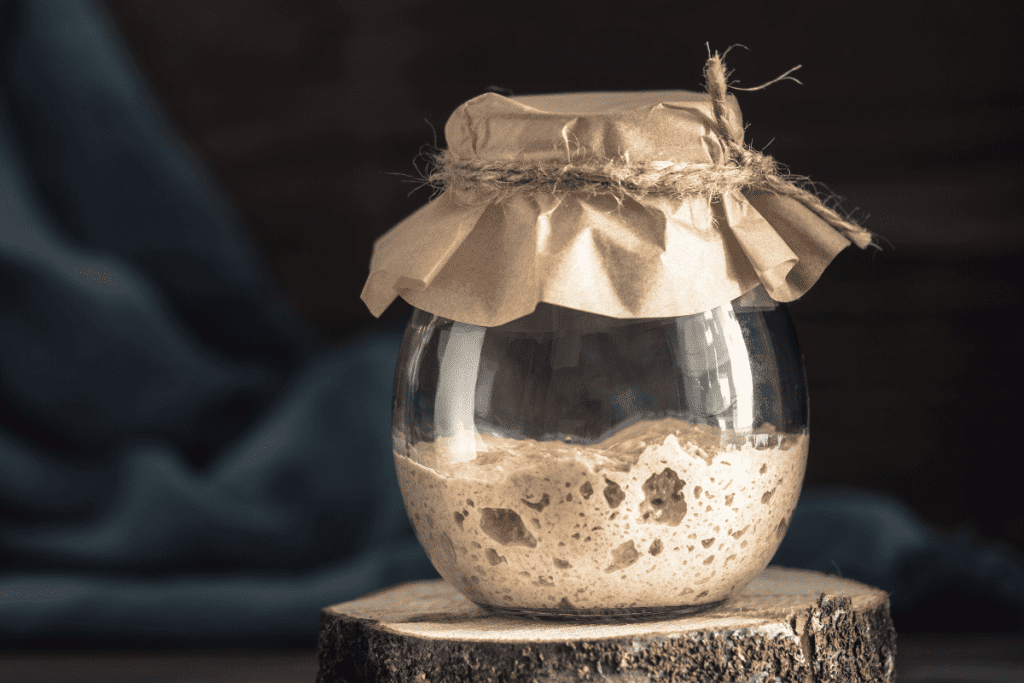 Sourdough Discard & Sourdough Starter | 100% Discard! {from Pantry Mama} 11 A partially filled glass jar of Sourdough Discard Recipes, starter, covered with kraft paper and tied with rustic twine, resting on a wood slice.