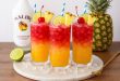 Tropical Malibu Bay Breeze cocktail in a highball glass with crushed ice, garnished with a maraschino cherry, lime slice, pineapple wedge, and a blue-striped straw on a marble board with a Malibu rum bottle and fresh pineapple in the background