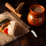 Traditional Mexican Rosca de Reyes slice with colorful candied fruit atop soft, sugar-dusted bread, paired with a rustic clay mug of hot chocolate and cinnamon sticks on a dark wooden table. A small baby figurine symbolizes the cultural tradition of Epiphany celebration.