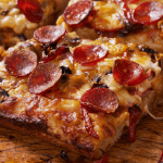 Close-up of a crispy Detroit-style pizza slice, highlighting the thick crust, caramelized cheese edges, and a delicious combination of toppings including pepperoni, black olives, and red bell peppers.
