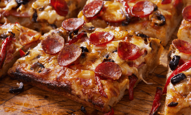 Close-up of a crispy Detroit-style pizza slice, highlighting the thick crust, caramelized cheese edges, and a delicious combination of toppings including pepperoni, black olives, and red bell peppers.