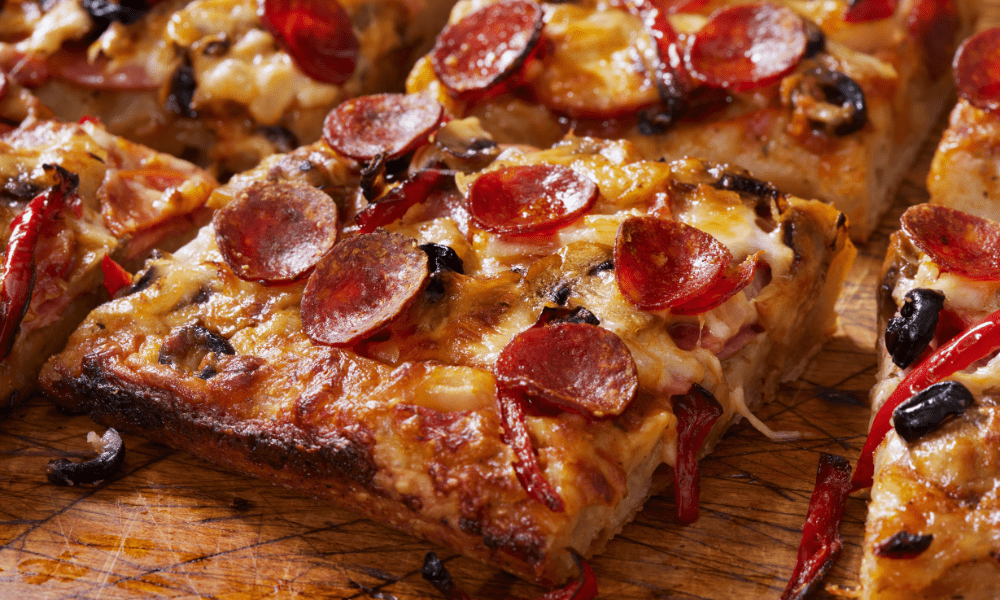 Close-up of a crispy Detroit-style pizza slice, highlighting the thick crust, caramelized cheese edges, and a delicious combination of toppings including pepperoni, black olives, and red bell peppers.