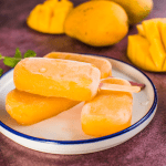 A close-up of vibrant yellow mango-flavored Mexican popsicles, or paletas, from La Michoacana, served on a plate with fresh mango slices and mint leaves, against a rustic background. yogurt