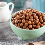 A mint green bowl filled with chocolate protein cereal, placed on a marble countertop alongside a white milk jug and a blue mug, perfect for a healthy breakfast setting