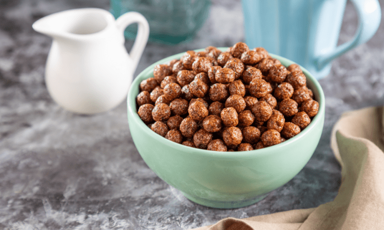 A mint green bowl filled with chocolate protein cereal, placed on a marble countertop alongside a white milk jug and a blue mug, perfect for a healthy breakfast setting