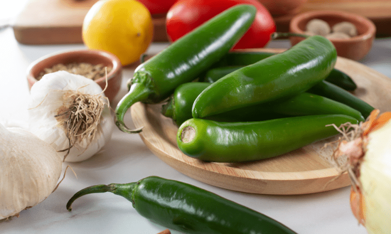 Fresh serrano peppers on a wooden plate surrounded by ingredients like garlic, lemon, and tomatoes, ready for cooking