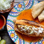A close-up of a traditional Mexican torta ahogada, soaked in spicy red sauce, garnished with pickled onions, and accompanied by lime and crispy tacos. A must-try in Mexican food culture.