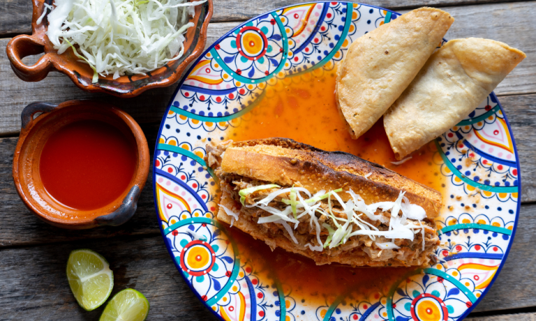 A close-up of a traditional Mexican torta ahogada, soaked in spicy red sauce, garnished with pickled onions, and accompanied by lime and crispy tacos. A must-try in Mexican food culture.