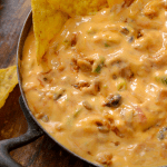 A close-up view of a thick, orange cheese dip filled with meat and chiles in a cast-iron skillet, surrounded by crunchy tortilla chips.