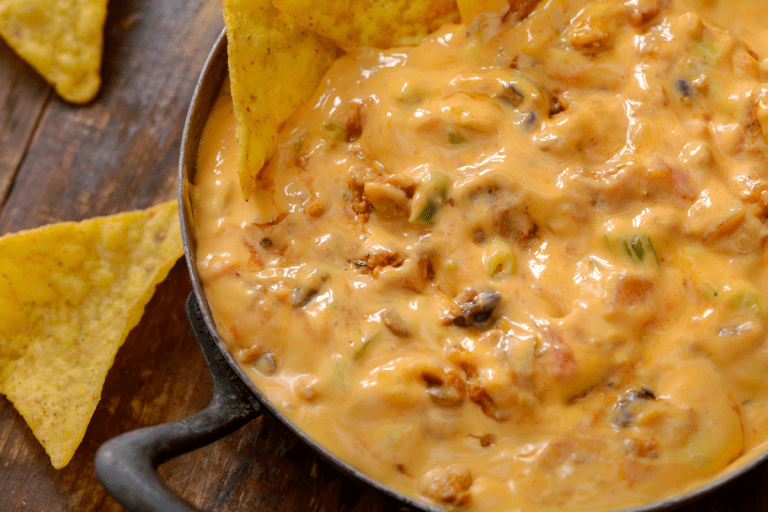 A close-up view of a thick, orange cheese dip filled with meat and chiles in a cast-iron skillet, surrounded by crunchy tortilla chips.