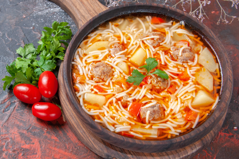 Top view of beef soup with meatballs, potatoes, and noodles, surrounded by fresh vegetables and spices on a dark table.
