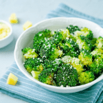 A simple white bowl packed with roasted broccoli, topped generously with grated cheese. The soft hues of the background make the green florets pop