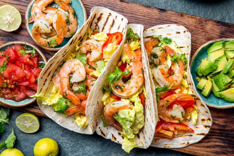 A closer look at shrimp cradled in soft tortillas and topped with chopped lettuce, peppers, and herbs. The accompanying bowls of pico de gallo and diced avocado invite you to customize each bite.