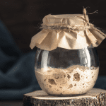 A partially filled glass jar of Sourdough Discard Recipes, starter, covered with kraft paper and tied with rustic twine, resting on a wood slice.