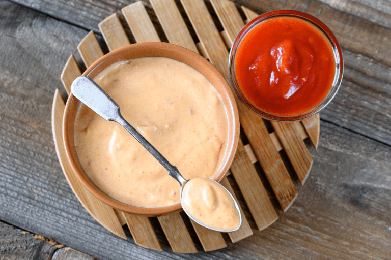 A single shot of a terracotta bowl filled with pink Yum Yum Sauce on a bamboo trivet, paired with a spoon and a small glass of ketchup on the side.