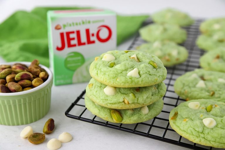 Stack of soft pistachio pudding cookies with white chocolate chips and chopped pistachios on a cooling rack, next to a box of pistachio pudding and a ramekin of whole pistachios